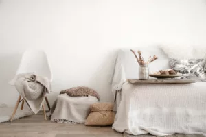 Styl wabi-sabi. Po lewej stronie widzmy krzesło z narzuconym na niego kocem. Obok krzesła stoi janobeżowa pufa z brązową poduszką. Obok niej stoi słomiany koszyk. W prawej części stoi łóżko - jego fragment. Na nim stoi tacka z wazonem i kwiatami oraz półmisek. Wszystko jest w barwach białych, beżowych i brązowych.