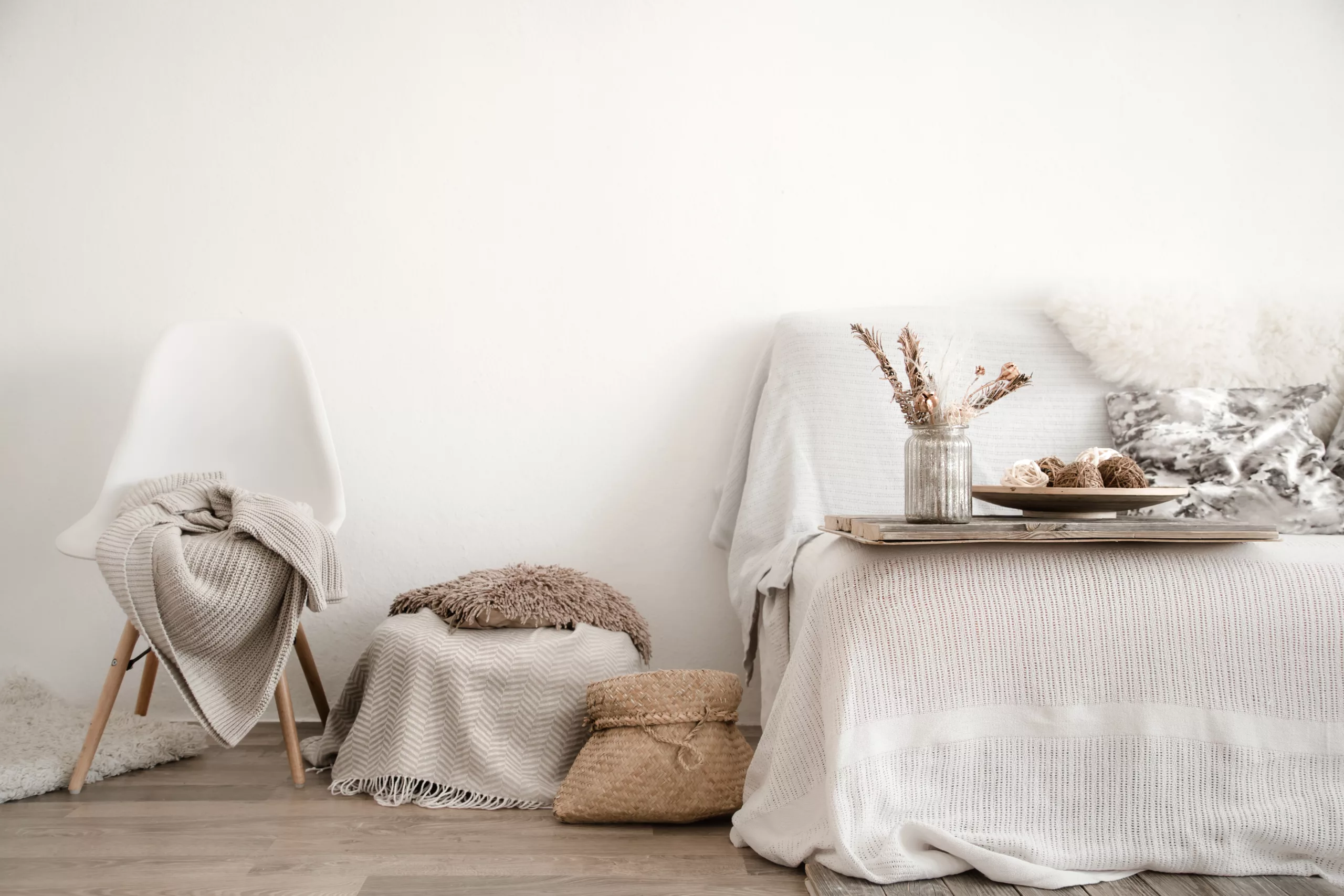 Styl wabi-sabi. Po lewej stronie widzmy krzesło z narzuconym na niego kocem. Obok krzesła stoi janobeżowa pufa z brązową poduszką. Obok niej stoi słomiany koszyk. W prawej części stoi łóżko - jego fragment. Na nim stoi tacka z wazonem i kwiatami oraz półmisek. Wszystko jest w barwach białych, beżowych i brązowych.